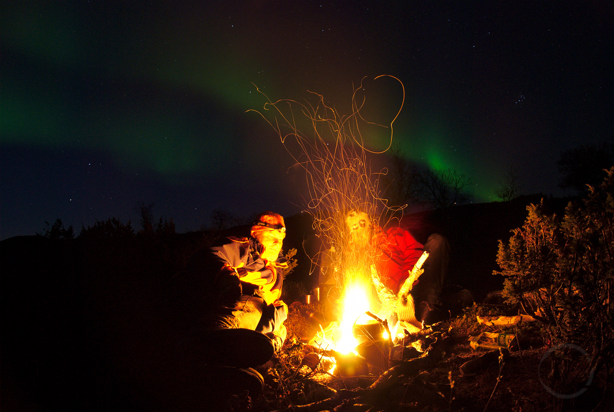 Two people sitting next two camp fire, with nothern lights in the background